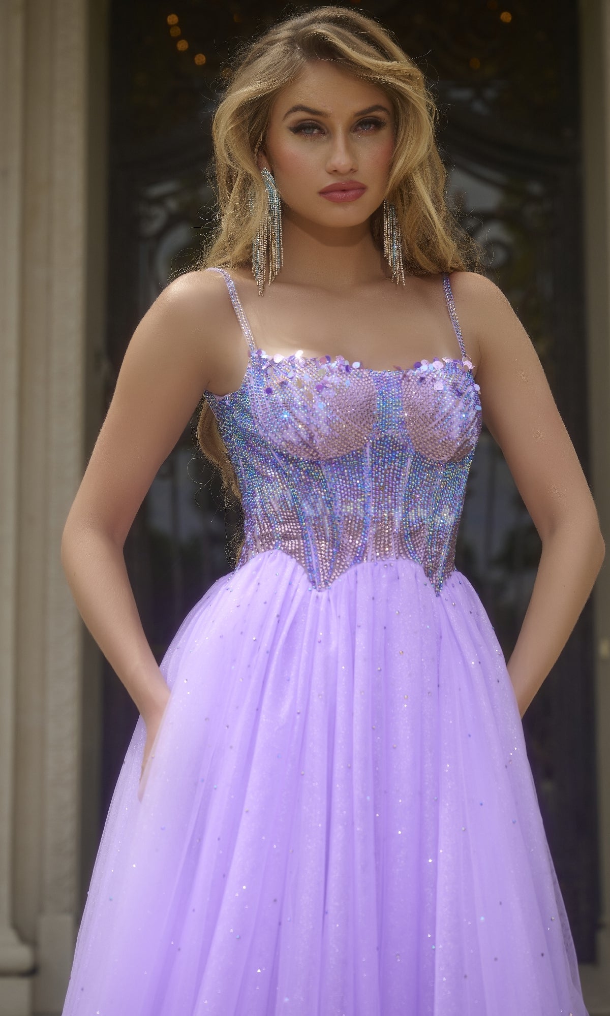 A woman stands in front of a building wearing the Jovani 44511 Beaded-Corset Long Prom Ball Gown with a tulle skirt, accessorized with long, dangling earrings.